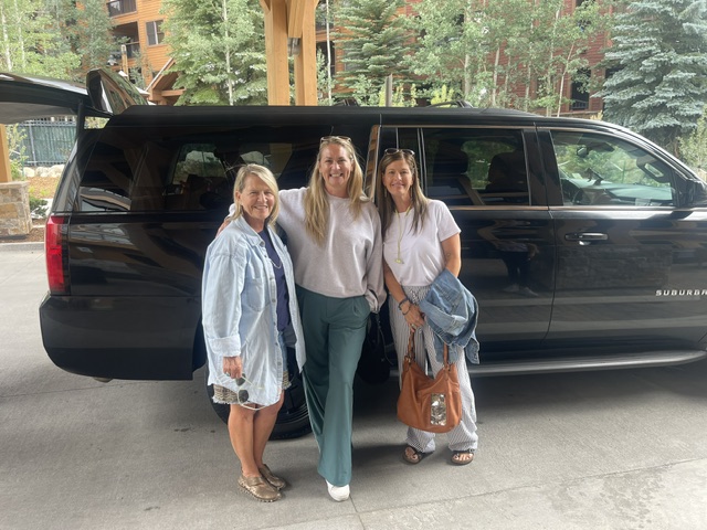 Three smiling travelers standing beside a black Suburban